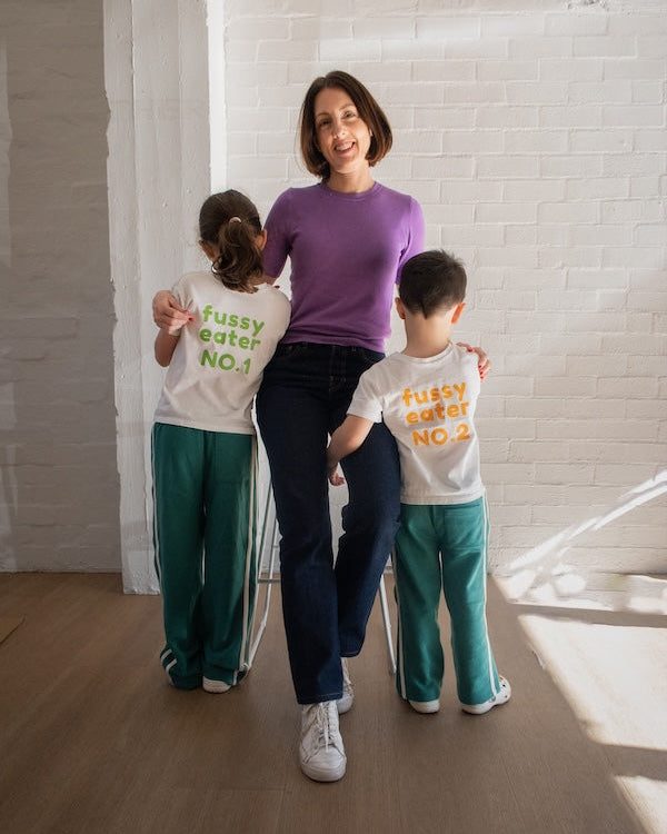 A woman stands between two children in a bright room with a white brick wall and wood floor. The woman wears a purple long-sleeve top and dark pants, while the children wear white t-shirts with colorful text and teal pants.