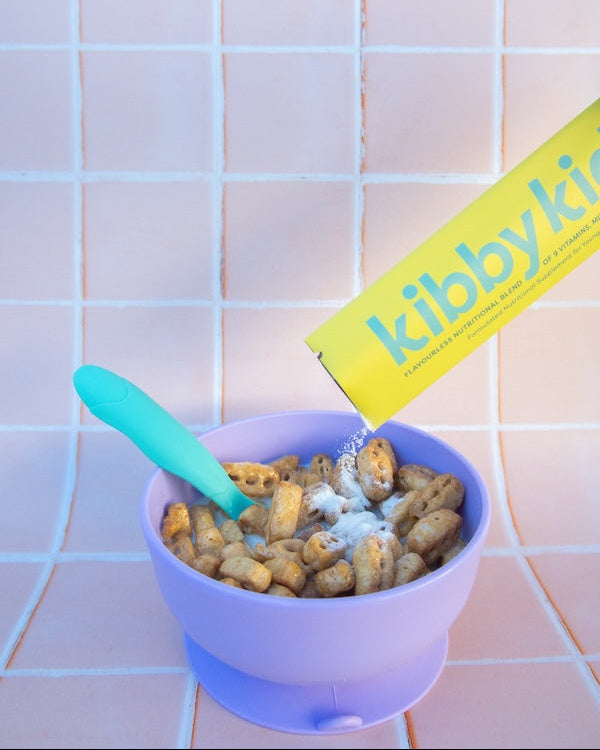 A yellow Kibby Kids cereal box pouring cereal into a lavender bowl with a turquoise spoon, set against a pink tiled background.