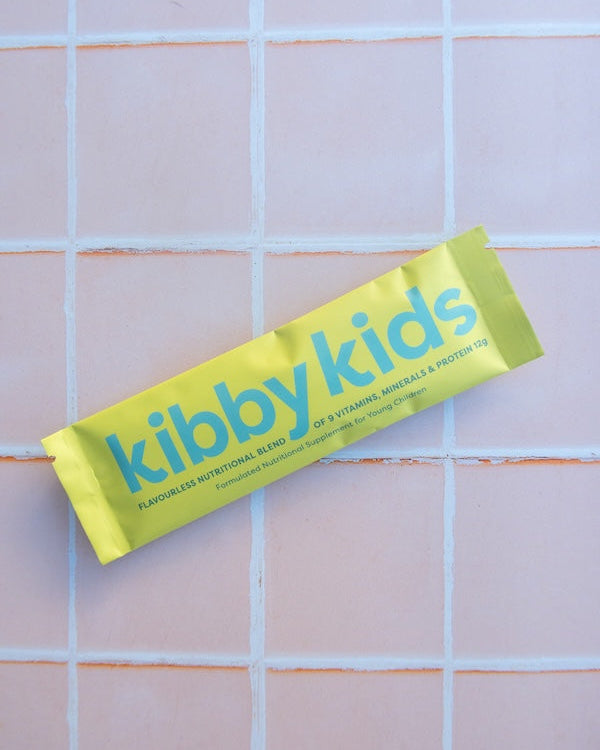A single kibby kids vitamin, mineral, and protein stick wrapped in a bright yellow-green foil wrapper, placed diagonally on a light pink tiled background.